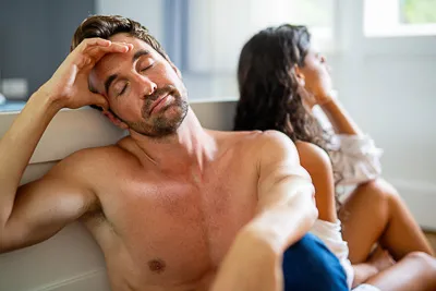 Hombre sin camisa con expresion de frustracion emocional, sentado junto a su pareja, simbolizando el impacto de la disfuncion erectil